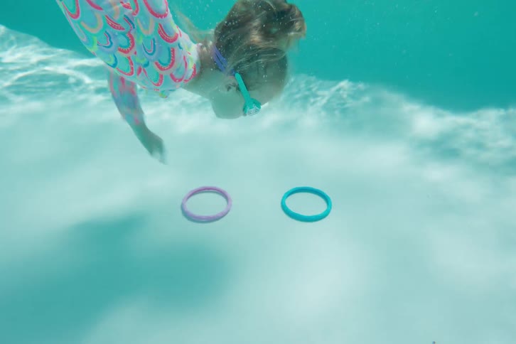 A young girl wearing goggles jumps into a swimming pool and dives underwater in slow motion to retrieve colorful toy rings from the bottom
