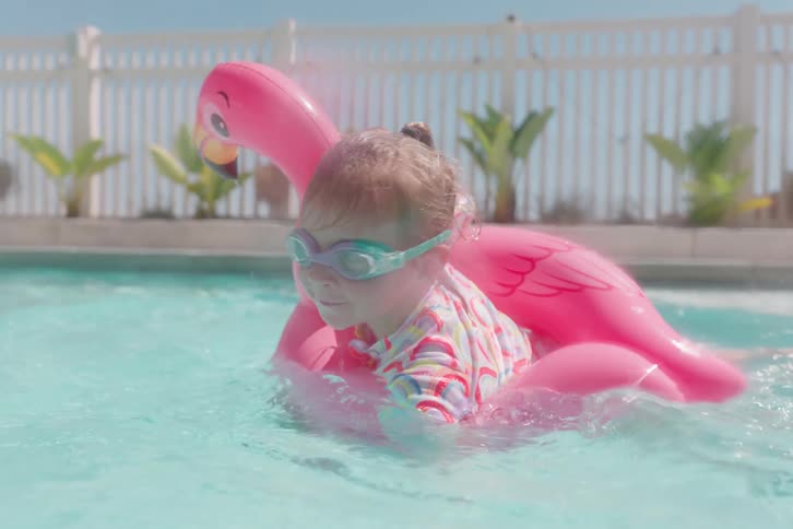 Happy little girl wearing goggles splashes into a backyard pool with her pink flamingo floaty on a sunny summer day in slow motion