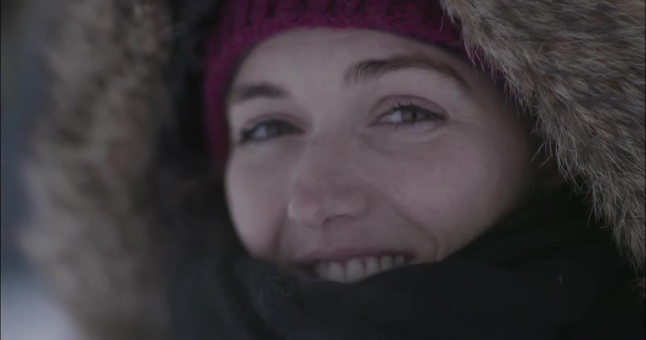 A woman smiles while outside in the cold dressed in a hooded heavy coat, skull cap, and a thick scarf