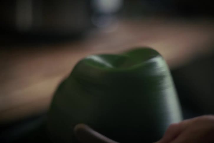 A hatter uses a brush to shape and finish a custom green felt hat as it spins on a block in a workshop