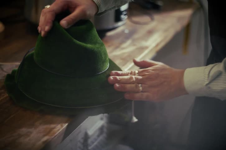 A skilled artisan meticulously brushes and shapes a traditional green velvet hat on a wooden workbench
