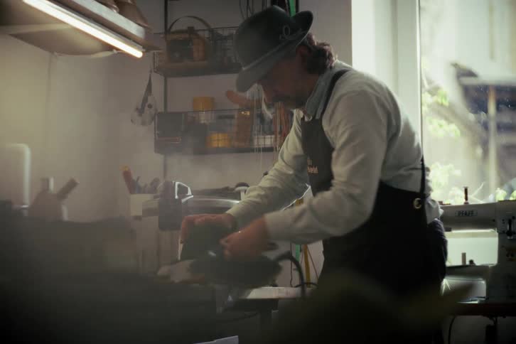 An artisan hatter meticulously irons the brim of a felt hat with steam in his cozy workshop