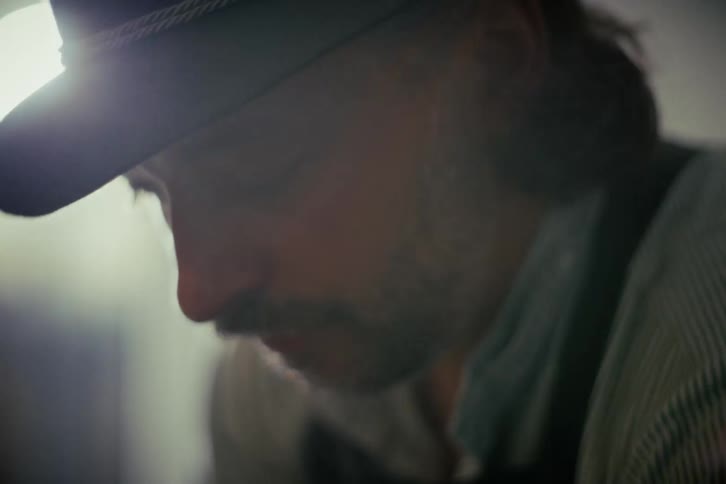 A moody, backlit close-up of a focused craftsman wearing a hat and sporting a mustache