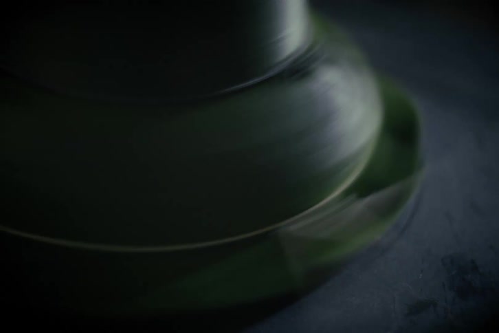 A hypnotic close-up of a green ceramic bowl spinning and wobbling on a potter's wheel in a dimly lit studio