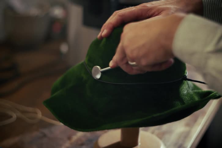 A craftsman uses a special tool to finish shaping a traditional green velvet hat in a workshop