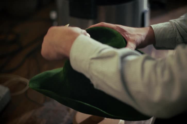 A hatter uses a wooden tool to meticulously shape a steamed green velvet hat in his workshop