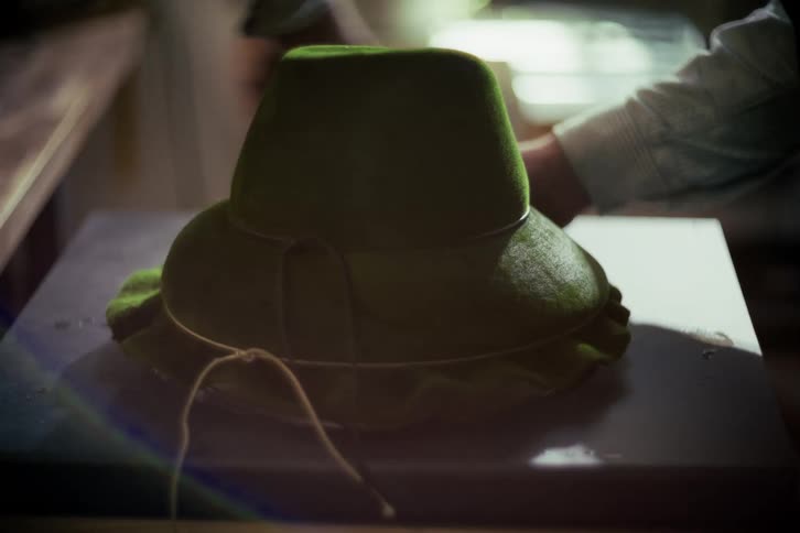 An artisan hat maker meticulously cleans and polishes a green felt hat in a sunlit workshop
