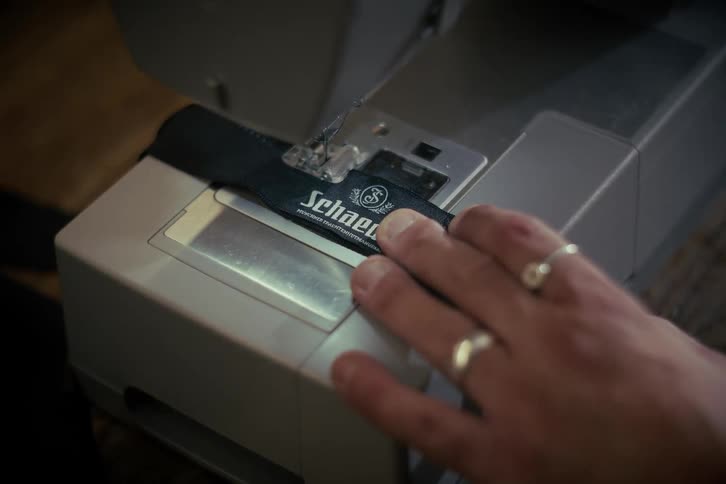 A detailed close-up of a person's hands using a sewing machine to attach a brand label to a piece of black fabric