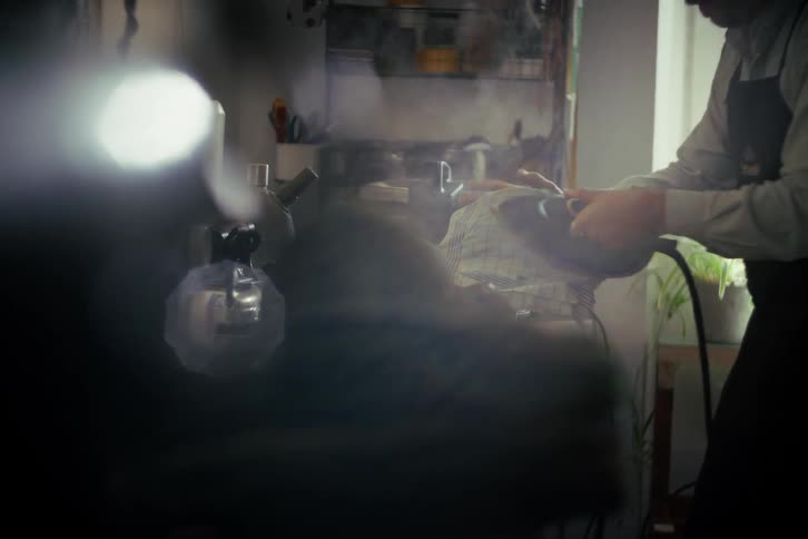 A craftsman in an apron meticulously irons and steams the brim of a hat in his artisan studio
