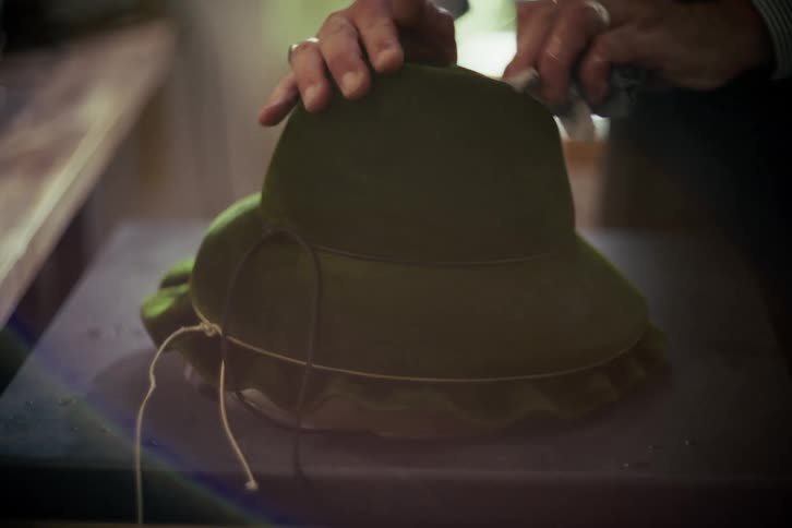Close-up of a hat maker's hands meticulously cleaning a green velvet hat on a block in a workshop