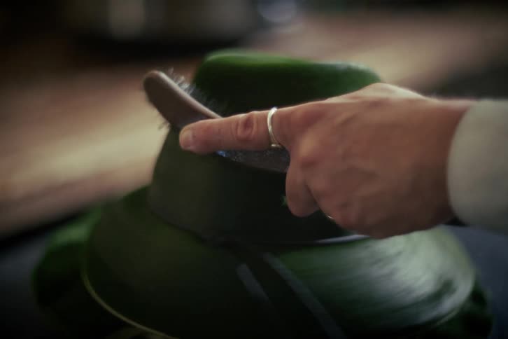 A craftsman meticulously brushes a traditional green fedora hat as it spins on a turntable in a workshop