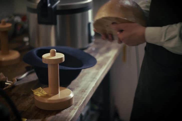 A craftsman removes a wooden block from the inside of a handmade hat and places it on a stand in a workshop