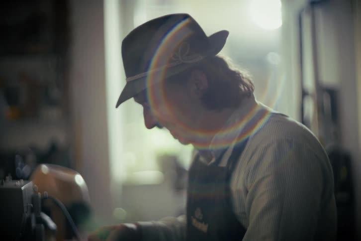 A mustachioed artisan in a traditional hat smiles and winks while working at his craft in a warmly lit workshop