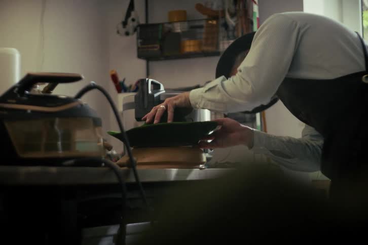 A milliner in an apron carefully molds a green felt hat over a wooden form in his workshop