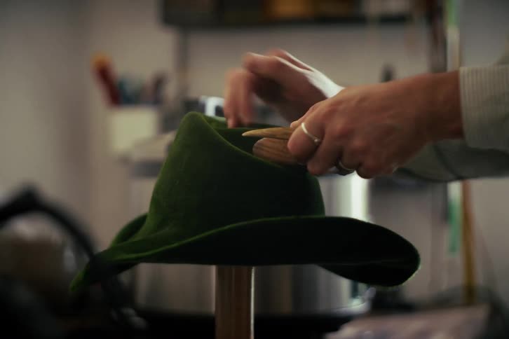 Close-up of an artisan meticulously brushing and shaping a green felt hat in their workshop