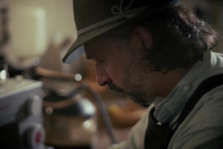 A craftsman with a long beard and a traditional hat concentrates on his work inside his workshop