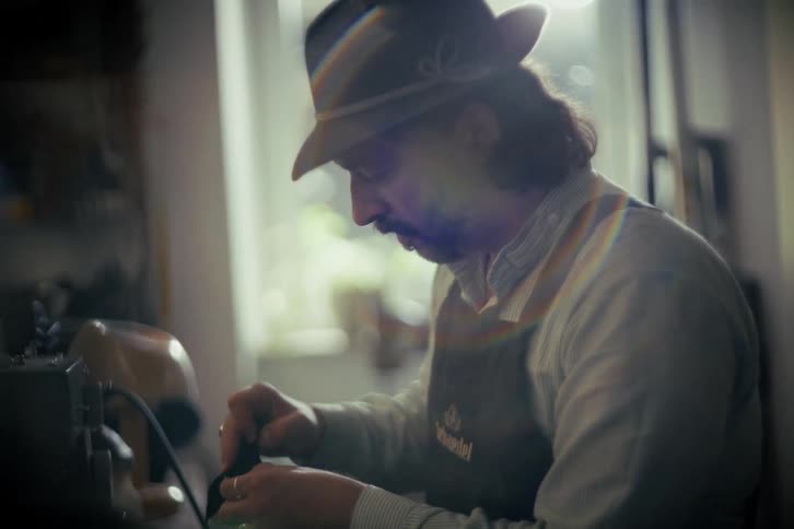 An artisan with a hat and apron carefully works on a piece of leather in his soft-lit, authentic workshop