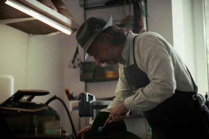 A skilled milliner meticulously shapes a traditional green felt hat with a specialized tool in his workshop