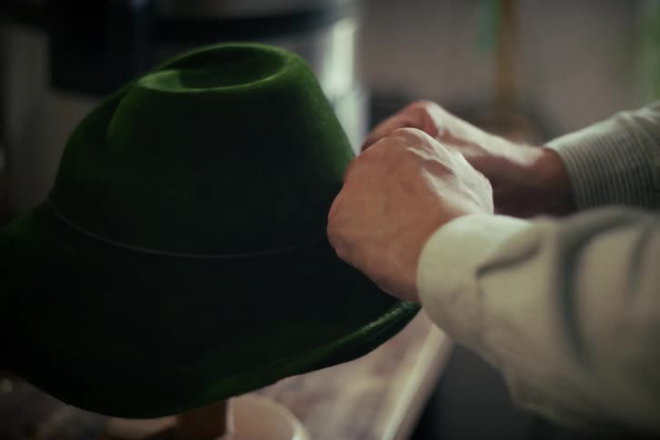 A skilled craftsman carefully ties a decorative cord band onto a traditional green felt hat in his workshop