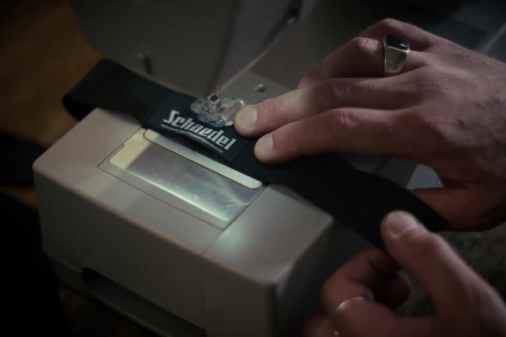 A close-up shot of a tailor's hands guiding a black fabric strap with a label through a sewing machine