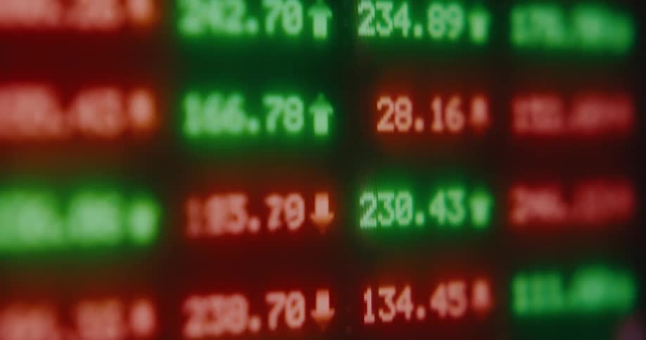 A blurry close-up of a digital stock market screen shows fluctuating green and red numbers indicating gains and losses
