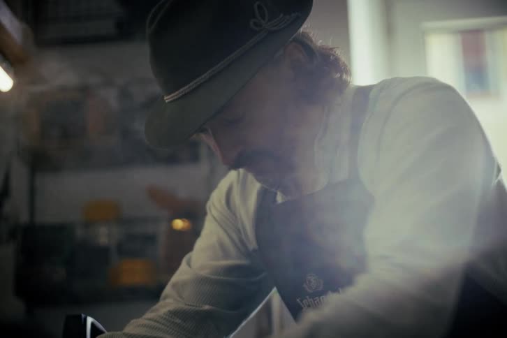 A traditionally dressed craftsman with a mustache and hat diligently works in his sunlit workshop as steam rises from his craft