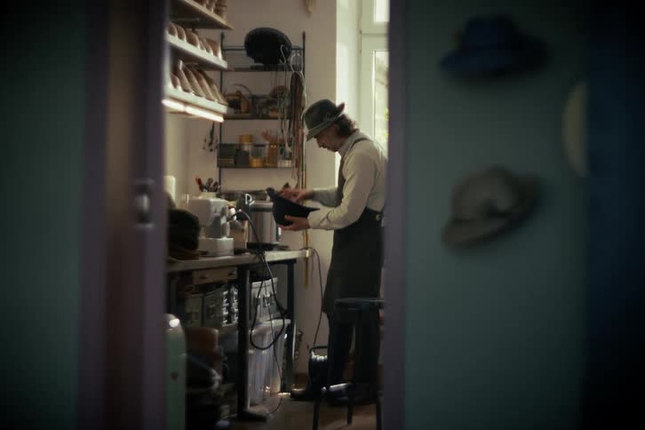 A craftsman wearing a fedora and apron carefully brushes and cleans the interior of a hat in his workshop