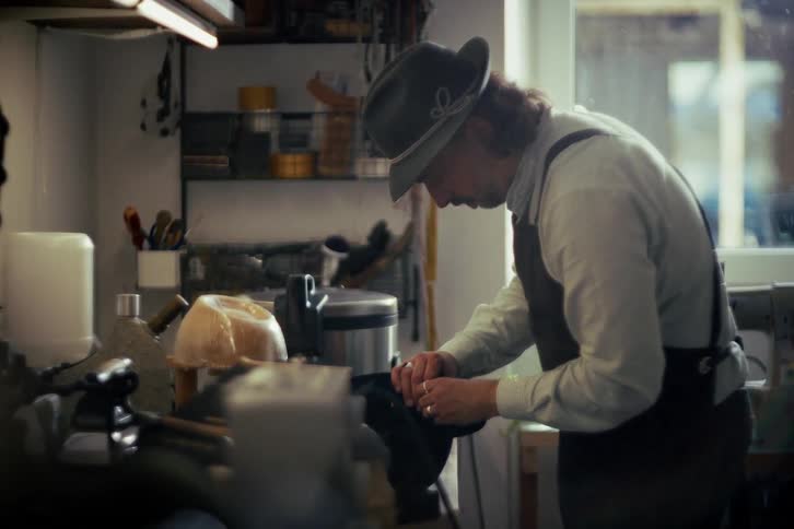 A skilled craftsman wearing a traditional hat meticulously works on a leather boot in his sunlit workshop