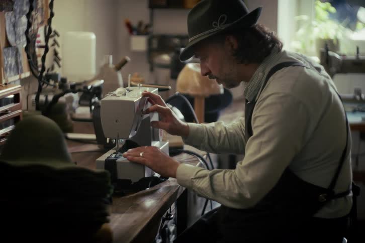 A traditional hat maker carefully sews a piece of fabric using a sewing machine in his rustic workshop