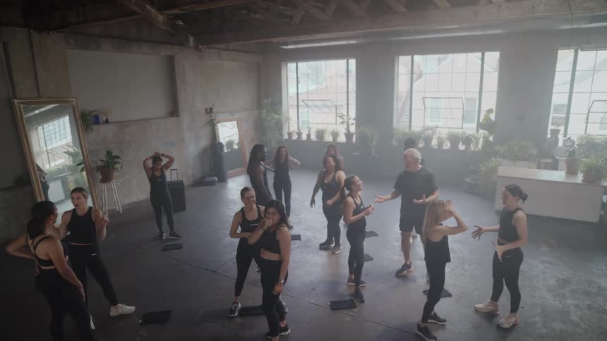 A diverse fitness class of mostly women chats and listens to their male instructor in a hazy, sunlit industrial loft studio