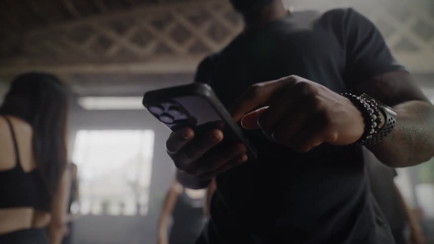 Close-up of a fit man in a black t-shirt using his smartphone inside a moody, industrial gym
