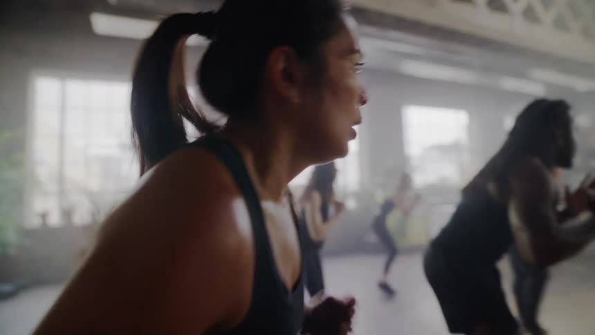 A focused woman sweats during a high-intensity group workout class in a hazy, industrial-style gym in slow motion