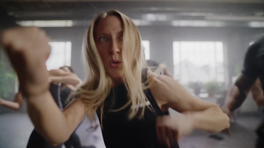 An intense woman with long blonde hair leads a group fitness class, throwing powerful punches towards the camera in slow motion