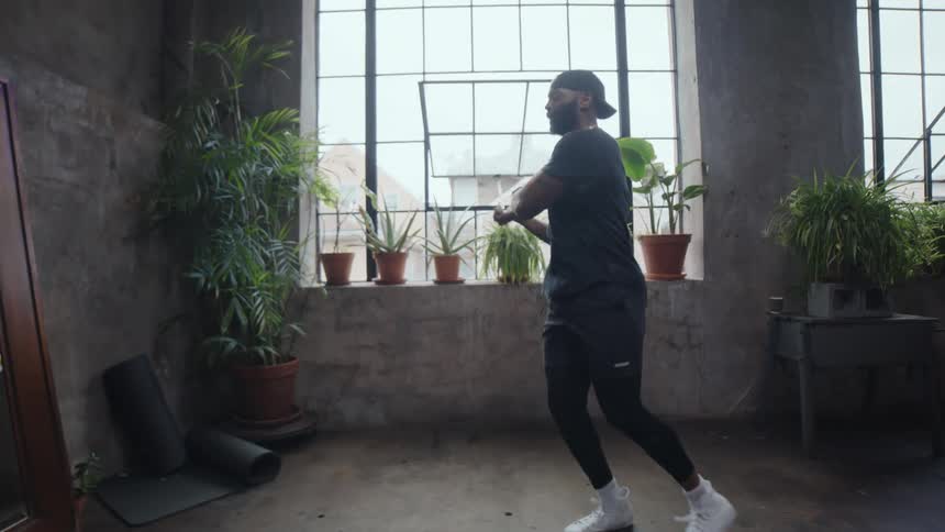 A fit man in workout gear jumps rope and uses a resistance band in slow motion inside a rustic home gym with large windows