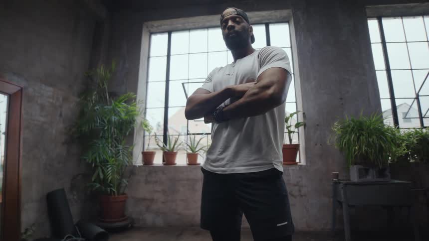 A determined athlete stands with arms crossed in a gritty, industrial gym as the camera slowly orbits around him
