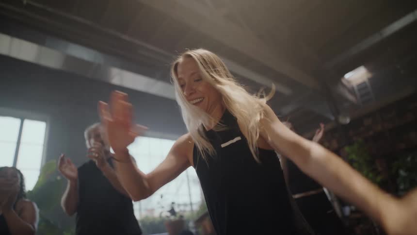 A joyful woman dances with abandon in a sunlit studio, cheered on by her friends in slow motion
