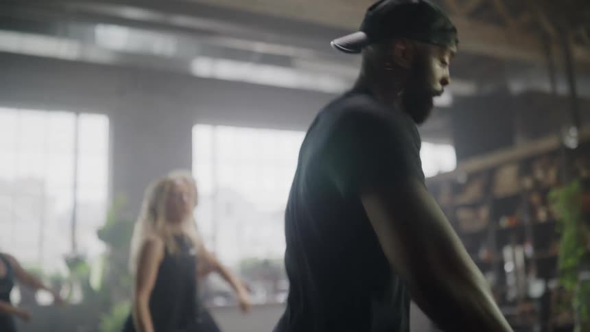 An energetic male instructor leads a diverse group of people in a fun, high-intensity dance fitness class in a sunlit studio