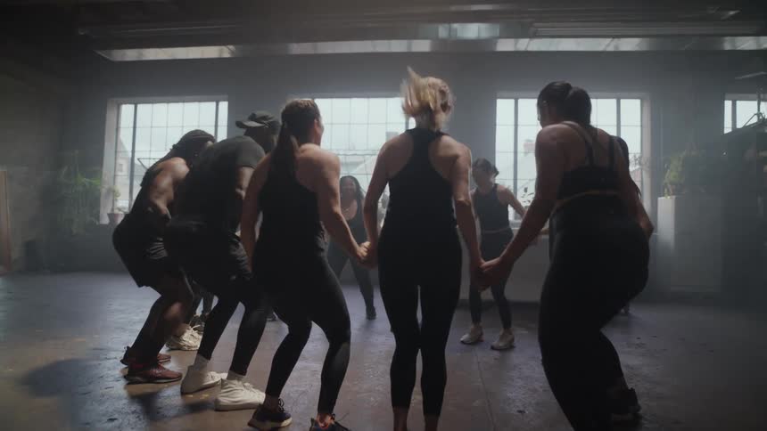 A diverse group of people hold hands in a circle and jump together during a high-energy workout class in a sunlit gym