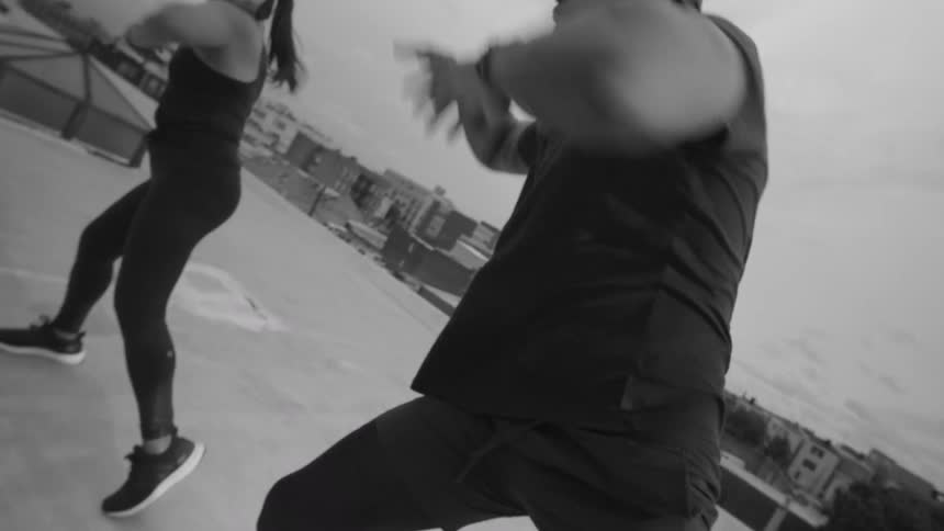 A man and woman perform a dynamic, synchronized workout on a city rooftop in a black and white slow-motion shot