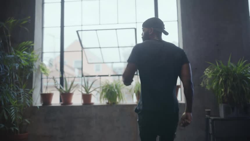 A man in a black t-shirt and backward baseball cap dances in slow motion in an industrial loft with large windows and potted plants