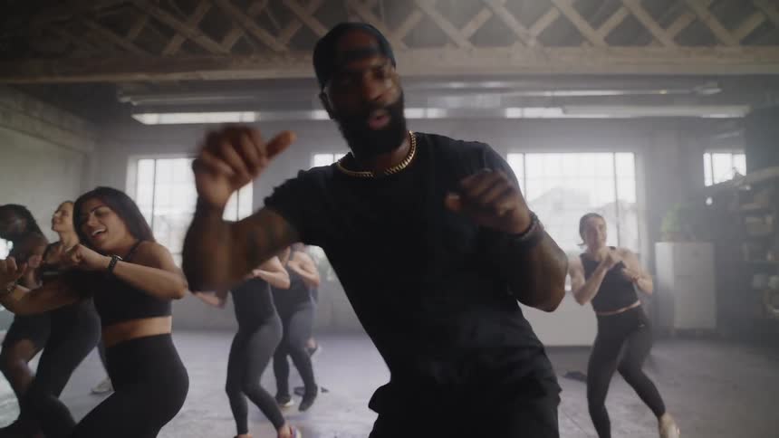 Slow motion shot of a charismatic male instructor leading a high-energy dance fitness class in a gritty, sunlit studio
