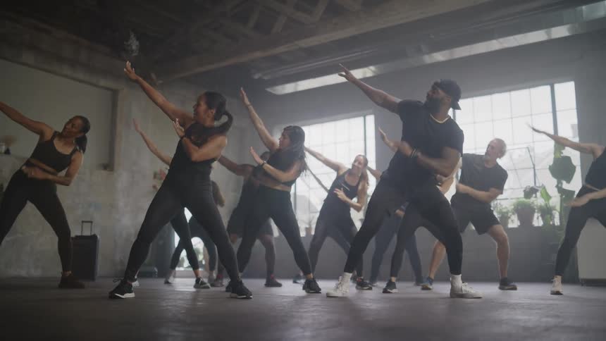 A diverse group of smiling dancers in black athletic wear follow their instructor in a high-energy fitness class in a hazy, sunlit studio