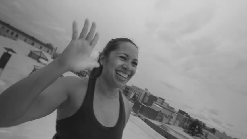 A joyful woman in workout clothes waves to the camera with a big smile in slow motion on an urban rooftop in black and white