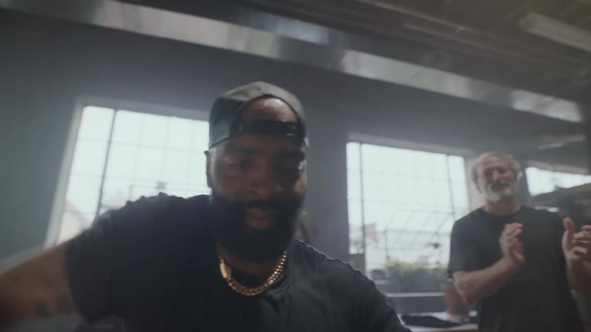A focused man leads a high-energy dance workout class, sweating profusely as his classmates enthusiastically clap and cheer him on in a hazy gym