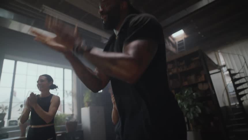 A muscular man gets hyped up by his diverse fitness class as they clap for him during a workout in a hazy, industrial-style gym