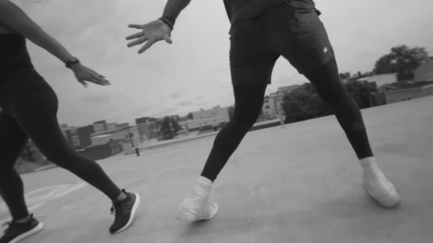 A smiling man and woman exercise together, doing synchronized squats on an urban rooftop in black and white slow motion