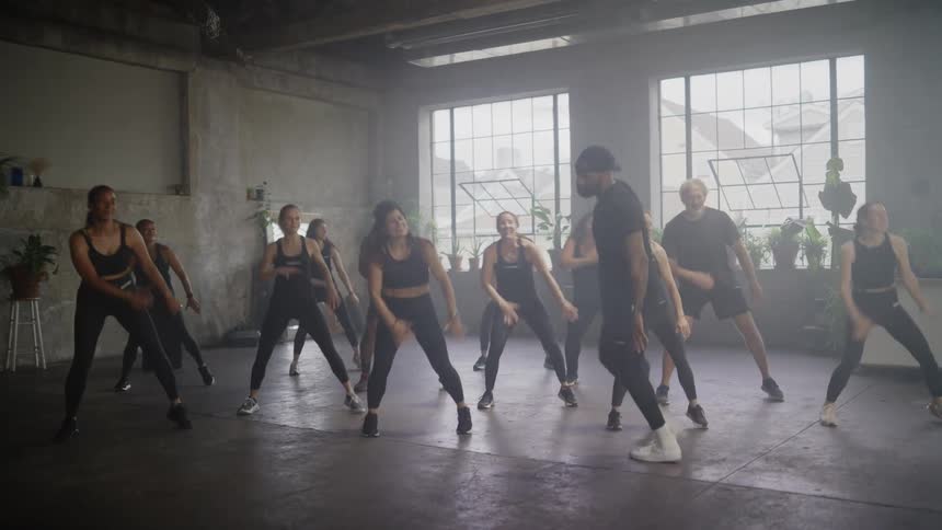 A diverse group of men and women energetically follow their instructor in a synchronized dance fitness class in a hazy, sunlit industrial studio
