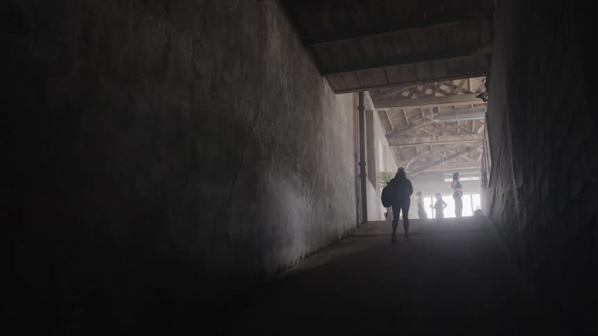 A silhouetted person carrying a gym bag walks up a dark ramp in slow motion towards a brightly lit exit