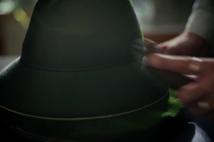 A close-up view of a person's hands carefully brushing and cleaning a classic dark green felt hat