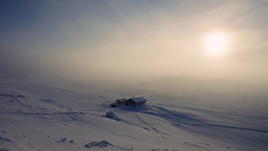 A remote wooden cabin sits alone in a vast, snowy landscape as the sun shines through the dense fog